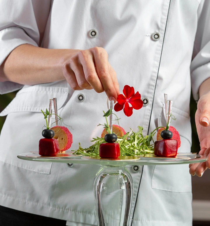 Chef plaatst een eetbare bloemengarnering op verfijnde canapéhapjes gerangschikt op een glazen serveerstandaard