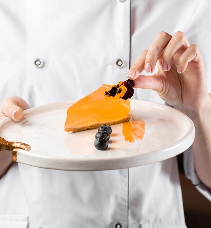 Chef plaatst een eetbare bloem op een stuk sinaasappeldessert geserveerd op een wit bord met blauwe bessen ter decoratie