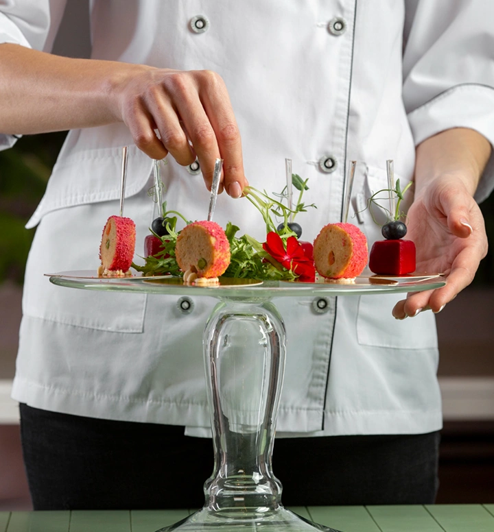 Chef presenteert elegante, hapklare gourmethapjes gerangschikt op een glazen serveerstandaard met kruiden en decoratieve toppings