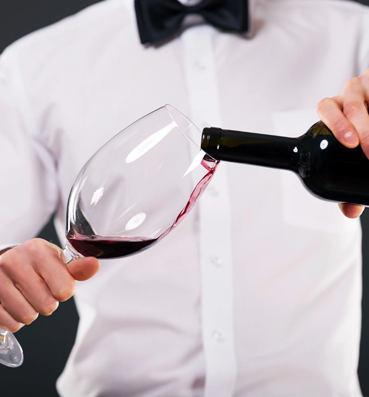 Ober in formeel uniform schenkt rode wijn in een groot glas tijdens een elegante dinerservice